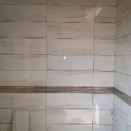 Ściana łazienki w trakcie układania płytek imitujących marmur, z dekoracyjnym pasem płytek w odcieniach brązu, widoczne krzyżyki dystansowe.