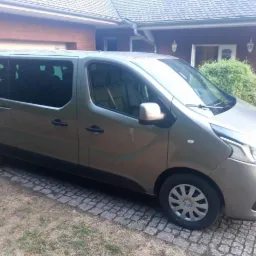 Szary bus Renault Trafic z przyciemnianymi szybami, zaparkowany na brukowanej nawierzchni przed domem z czerwonej cegły.