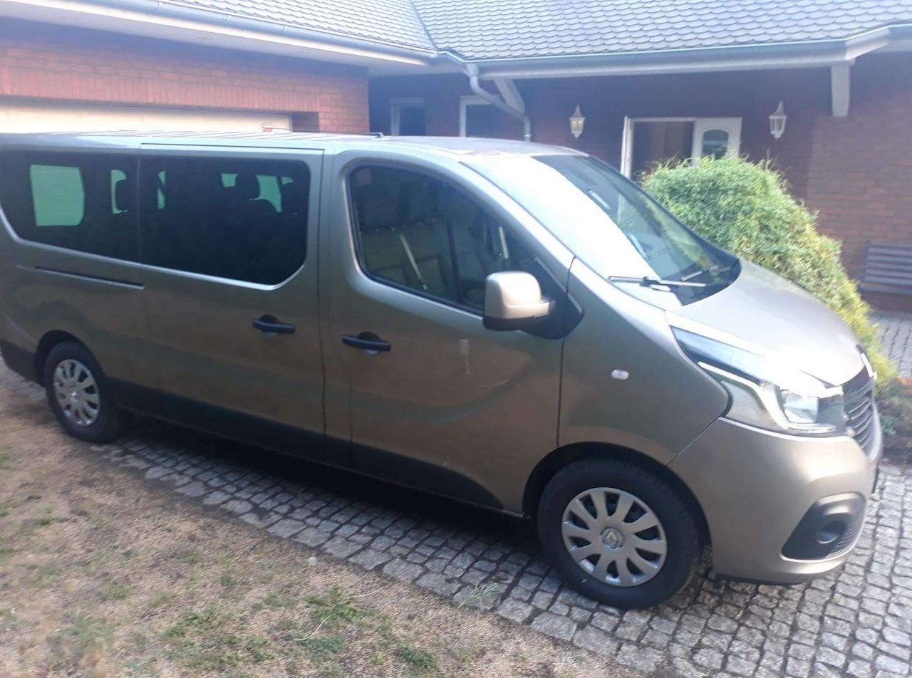 Szary bus Renault Trafic z przyciemnianymi szybami, zaparkowany na brukowanej nawierzchni przed domem z czerwonej cegły.