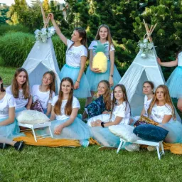 Grupa dziewcząt w błękitnych tiulowych spódniczkach i białych koszulkach z logo siedzi na trawie przed dwoma białymi tipi ozdobionymi kwiatami, jedna dziewczyna trzyma pluszowego ananasa.