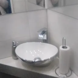 Nowoczesna, okrągła umywalka nablatowa z chromowanym wykończeniem, srebrny dozownik mydła i biały uchwyt na papier toaletowy na tle białych płytek i lustra z odbiciem osoby w białej kurtce.