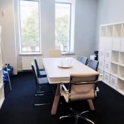 Jasna sala konferencyjna z długim stołem, krzesłami, regałami i oknami z widokiem na zieleń, na stole szklana karafka i szklanki.