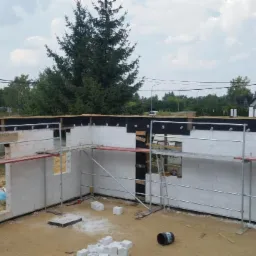 Wznoszenie ścian parteru domu jednorodzinnego z białych bloczków, widoczne rusztowania, mężczyzna na rusztowaniu robiący zdjęcia, w tle dom sąsiada i zieleń