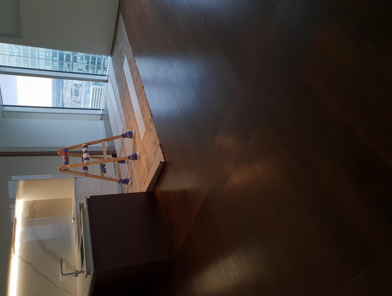 Ciemny parkiet ułożony we wzór jodełki francuskiej, częściowo zakryty płytami przygotowanymi do montażu, z widoczną drewnianą drabinką z niebieskimi zabezpieczeniami na nogach, w tle okno...