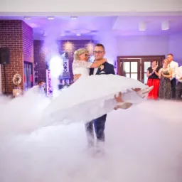 Pan młody trzyma pannę młodą na rękach podczas pierwszego tańca w chmurach, goście weselni w tle.