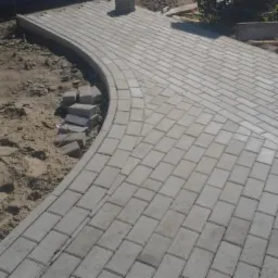 Fragment chodnika z szarej kostki brukowej w trakcie budowy, widoczne niedokończone obrzeże i przygotowane kostki obok piaszczystego podłoża.