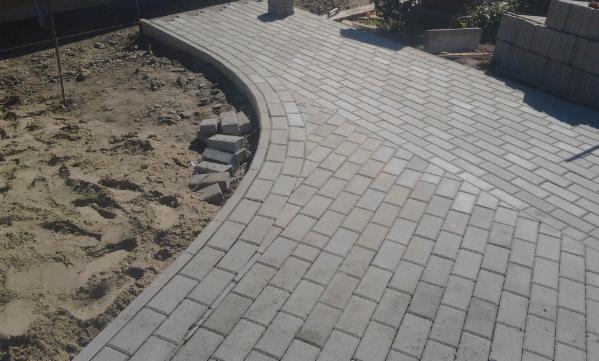 Fragment chodnika z szarej kostki brukowej w trakcie budowy, widoczne niedokończone obrzeże i przygotowane kostki obok piaszczystego podłoża.