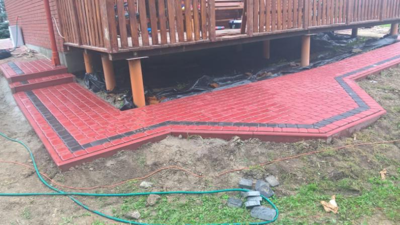 Czerwona kostka brukowa z czarnymi akcentami prowadząca od schodów do podwyższonego tarasu, widoczne rury wspierające taras i przygotowany teren pod dalsze prace.