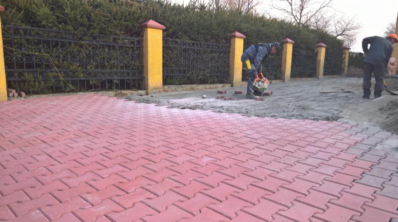 Układanie czerwonej kostki brukowej typu 'jodełka' na posesji z żółtymi słupkami ogrodzeniowymi; pracownik docina kostkę szlifierką kątową.