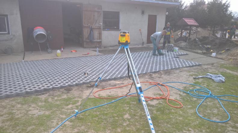 Montaż ażurowych płyt chodnikowych na posesji, widoczny niwelator laserowy na trójnogu, betoniarka i pracownicy w tle.