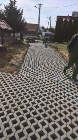 Nowo ułożona, ażurowa nawierzchnia z betonowych elementów w kształcie rombów, z widoczną linią wytyczającą, w tle dom jednorodzinny i prace ziemne.