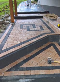 Ułożona kostka brukowa w geometryczny wzór z ciemnymi obramowaniami, prowadząca do schodów wejściowych domu. Widoczny gumowy młotek brukarski.