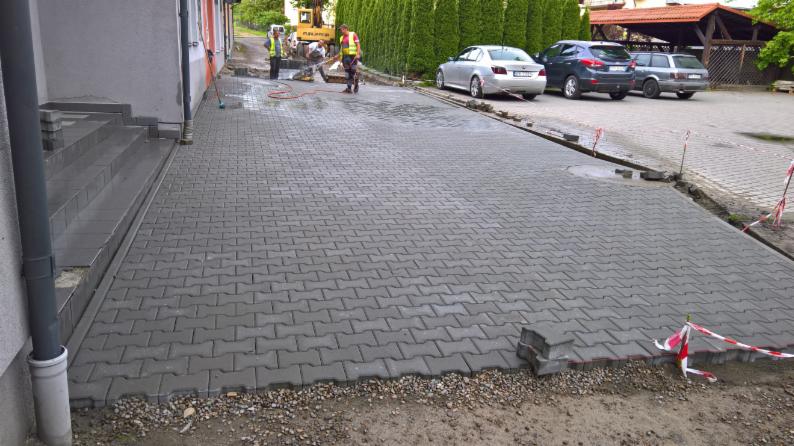 Układanie szarej kostki brukowej typu 'I' na posesji, widoczni pracownicy w odblaskowych kamizelkach i sprzęt budowlany w tle, częściowo ukończona nawierzchnia z elementami zabezpieczającymi.