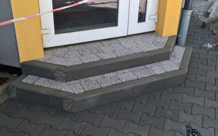 Trzy stopnie wejściowe wyłożone szarą kostką granitową, obrzeża z ciemnoszarej kostki, widoczne fragmenty elewacji budynku i kostki brukowej na chodniku.
