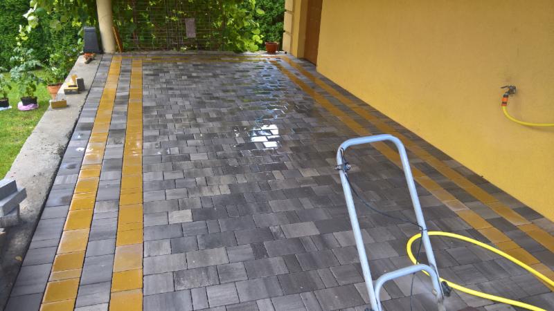 Świeżo ułożona, mokra kostka brukowa w odcieniach szarości z dwoma pasami żółtej kostki, myta myjką ciśnieniową przy żółtej ścianie budynku i zieleni.