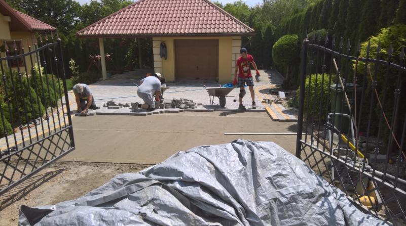 Prace brukarskie w toku: dwóch mężczyzn układa kostkę na podjeździe, widoczne narzędzia, taczka i plandeka zakrywająca materiał. W tle brama wjazdowa, dom z garażem i bujna roślinność.