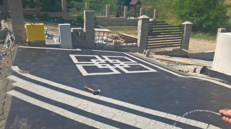 Nowo ułożony podjazd z ciemnej kostki brukowej z białymi pasami i dekoracyjnym wzorem w kształcie kwadratu, widoczny gumowy młotek i częściowo wykonane ogrodzenie z kamiennymi słupkami.