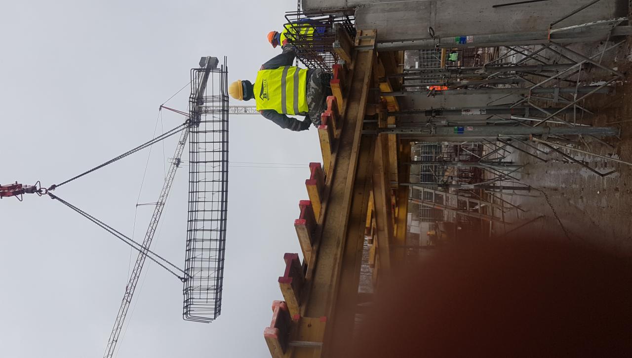 Pracownicy w żółtych kamizelkach montują zbrojenie konstrukcji żelbetowej przy użyciu dźwigu, widoczne szalunki i podpory.