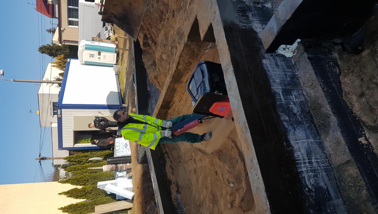 Pracownik w żółtej kamizelce zagęszcza grunt w wykopie pod fundamenty za pomocą zagęszczarki wibracyjnej; widoczna izolacja pionowa fundamentów i kontener biurowy w tle.