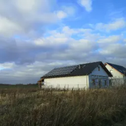 Nowe domy jednorodzinne z panelami słonecznymi na dachu, widoczne rusztowania przy jednym z budynków, w tle pochmurne niebo i pole z suchą trawą.