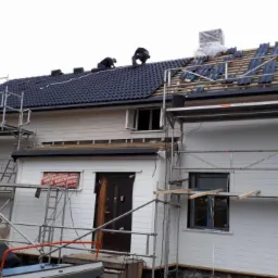 Dwóch dekarzy pracuje na dachu domu jednorodzinnego, montując ciemną dachówkę; widoczne rusztowania, częściowo pokryty dach i przygotowane elementy dachówki.