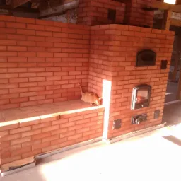 Nowo wybudowany piec kaflowy z cegły z pomarańczowym kotem jedzącym karmę na półce, w jasnym słońcu.