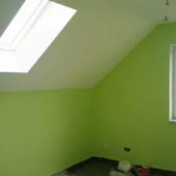 Pomalowane na jasnozielono poddasze z oknem dachowym i grzejnikiem pod oknem, widoczne niedociągnięcia w wykończeniu podłogi.