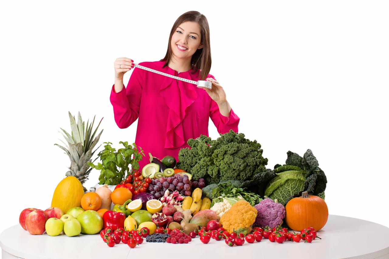 Uśmiechnięta kobieta w różowej bluzce trzyma miarkę krawiecką na tle stołu z różnorodnymi owocami i warzywami, w tym ananasem, dynią, brokułami i pomidorami koktajlowymi.