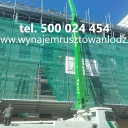 AD-BUD Rusztowania Wynajem Łódź - Elewacja budynku w rusztowaniach, zabezpieczona siatką. Na pierwszym planie wysięgnik pompy do betonu. Widoczny pracownik na rusztowaniu.