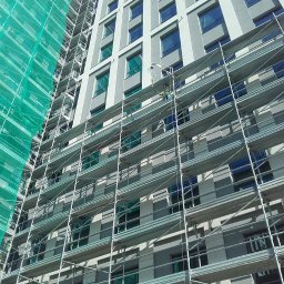 AD-BUD Rusztowania Wynajem Łódź - Wysoki budynek w trakcie prac elewacyjnych, osłonięty rusztowaniami i siatką ochronną. Widoczny pracownik na rusztowaniu. Ujęcie z dołu, podkreślające wysokość budynku.