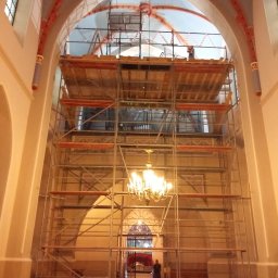 AD-BUD Rusztowania Wynajem Łódź - Wnętrze kościoła z rusztowaniem, widoczne prace na sklepieniu. Żyrandol i ławki w dolnej części kadru. Osoba w oddali przy wejściu.