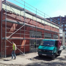 AD-BUD Rusztowania Wynajem Łódź - Budynek w trakcie elewacji z cegły, rusztowania, pracownik w kamizelce odblaskowej i zielony samochód dostawczy z przyczepą na placu budowy.