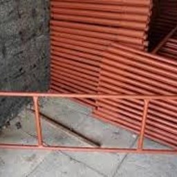 AD-BUD Rusztowania Wynajem Łódź - Pomarańczowe, metalowe rusztowanie modułowe na tle szarej ściany i stosu rur. Widoczne elementy konstrukcyjne, złącza i charakterystyczna geometria rusztowania.