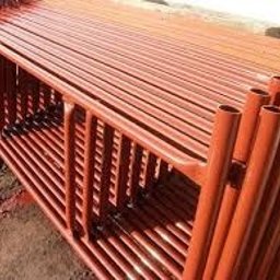 AD-BUD Rusztowania Wynajem Łódź - Rząd pomarańczowych, metalowych rusztowań budowlanych ułożonych jeden na drugim, widocznych pod kątem, z fragmentem ściany w tle.