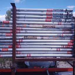 AD-BUD Rusztowania Wynajem Łódź - Stos rur rusztowania Boels Rental, ułożonych poziomo na przyczepie. Widoczne ślady użytkowania i naklejki firmowe. Tło: niebieskie niebo z chmurami i zieleń drzew.