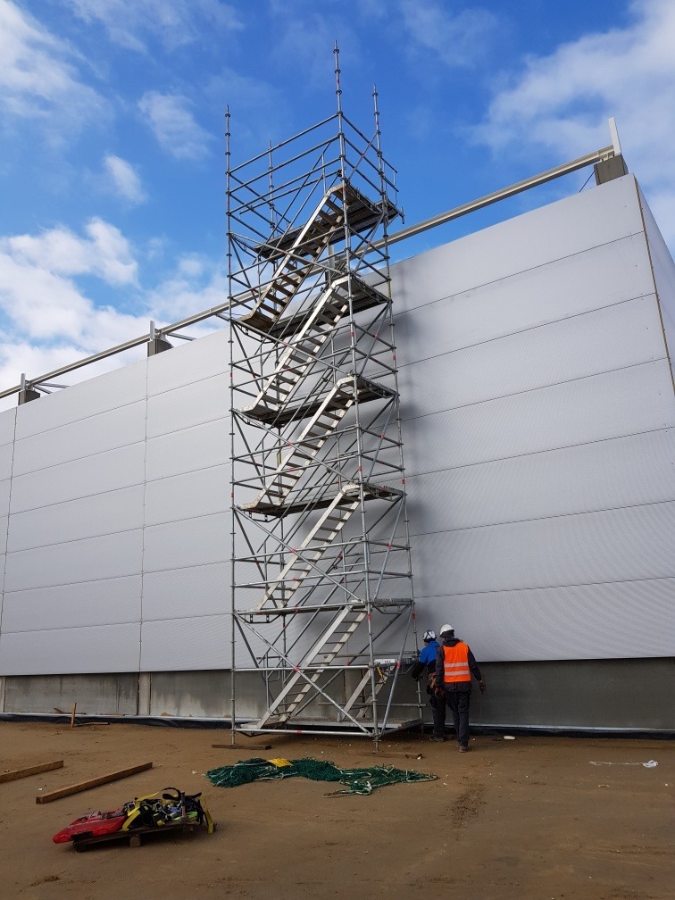Wysoka wieża rusztowania schodowego przy elewacji budynku przemysłowego, dwóch pracowników w odblaskowych kamizelkach przy podstawie, błękitne niebo w tle.