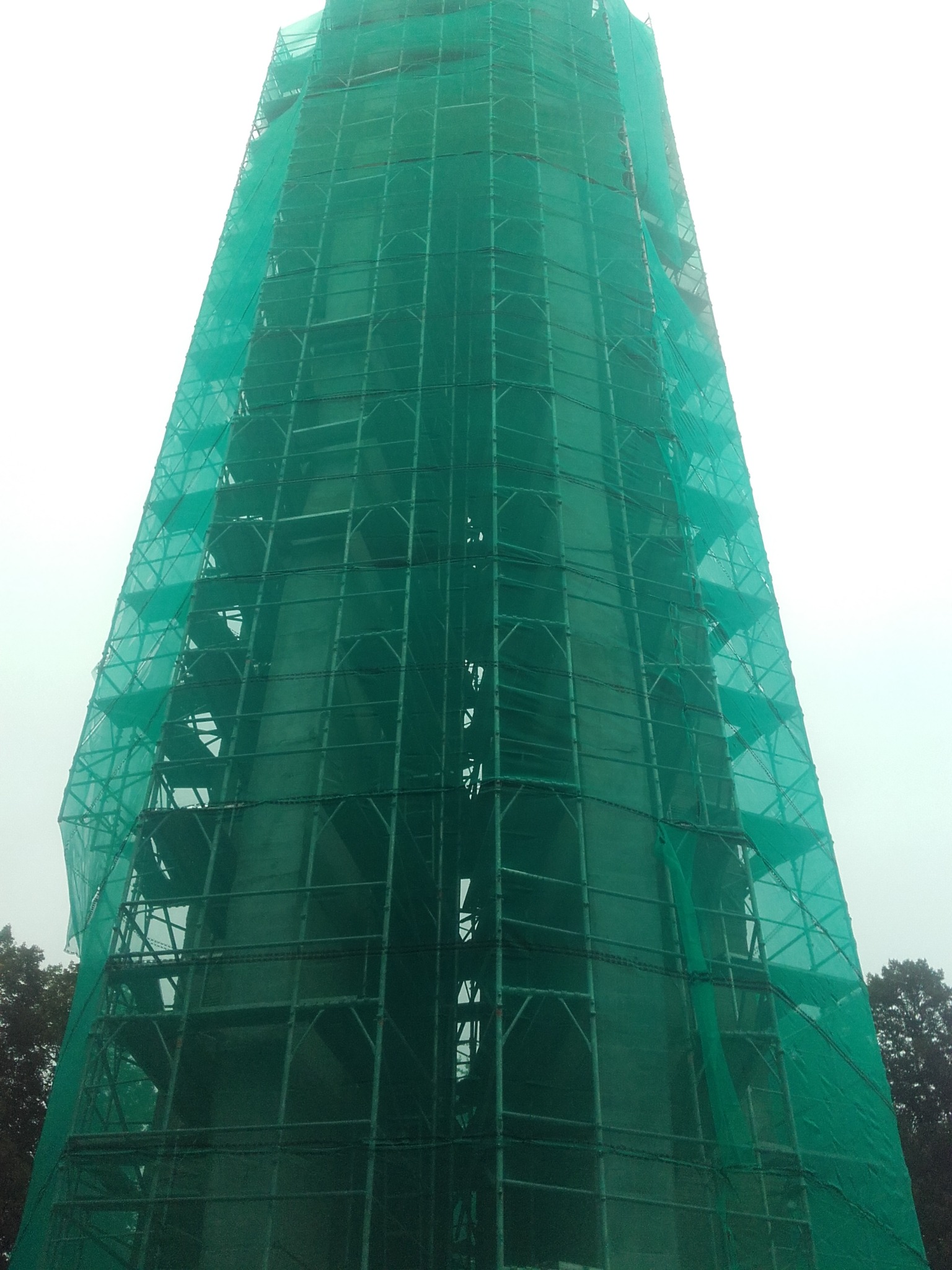 Wieża w rusztowaniach, osłonięta zieloną siatką ochronną, widoczna z dołu. Prace budowlane w toku, drzewa w tle. Mglista pogoda.