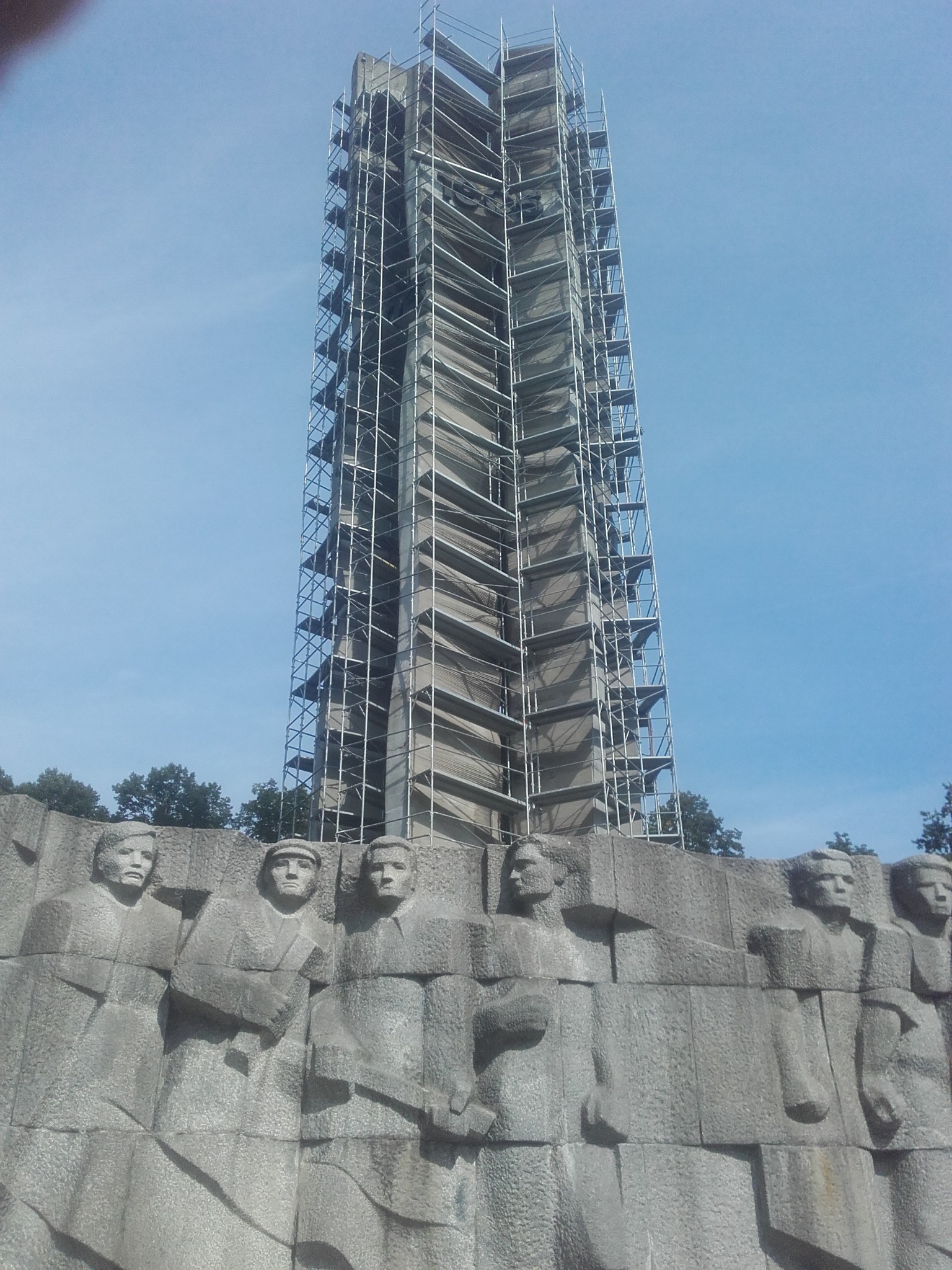 Wysoka, kamienna kolumna otoczona rusztowaniem, częściowo zakryta siatką ochronną. U dołu płaskorzeźba z postaciami. Błękitne niebo w tle.