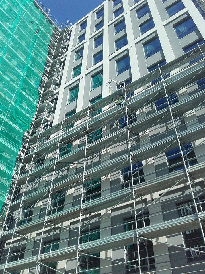 Wysoki budynek w trakcie prac elewacyjnych, osłonięty rusztowaniami i siatką ochronną. Widoczny pracownik na rusztowaniu. Ujęcie z dołu, podkreślające wysokość budynku.