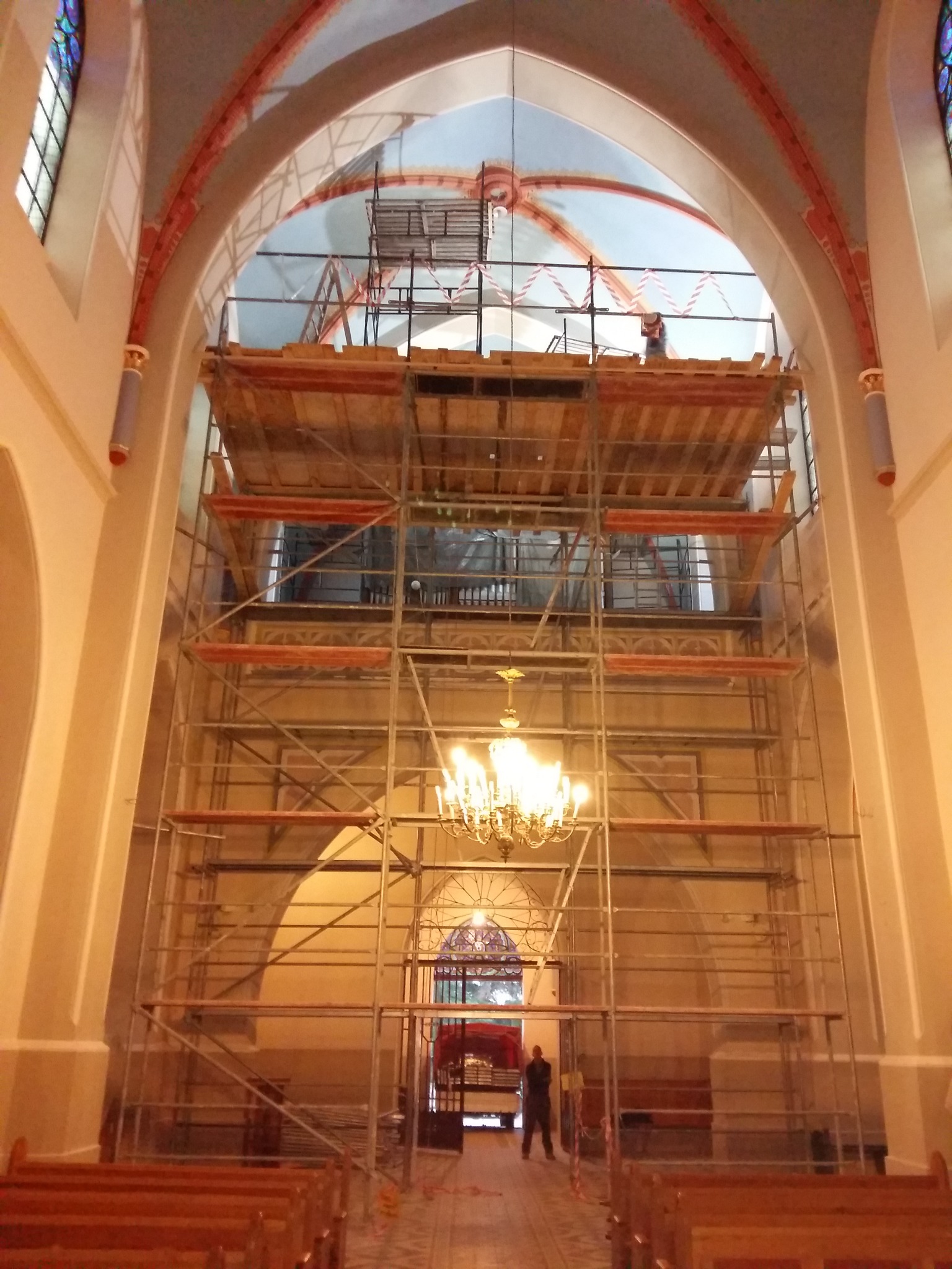 Wnętrze kościoła z rusztowaniem, widoczne prace na sklepieniu. Żyrandol i ławki w dolnej części kadru. Osoba w oddali przy wejściu.