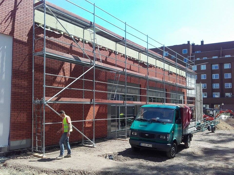 Budynek w trakcie elewacji z cegły, rusztowania, pracownik w kamizelce odblaskowej i zielony samochód dostawczy z przyczepą na placu budowy.