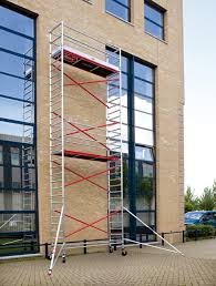 Aluminiowe rusztowanie wieżowe na kółkach, ustawione przy elewacji budynku z dużymi oknami. Widoczne czerwone elementy konstrukcyjne i beżowa cegła elewacyjna.