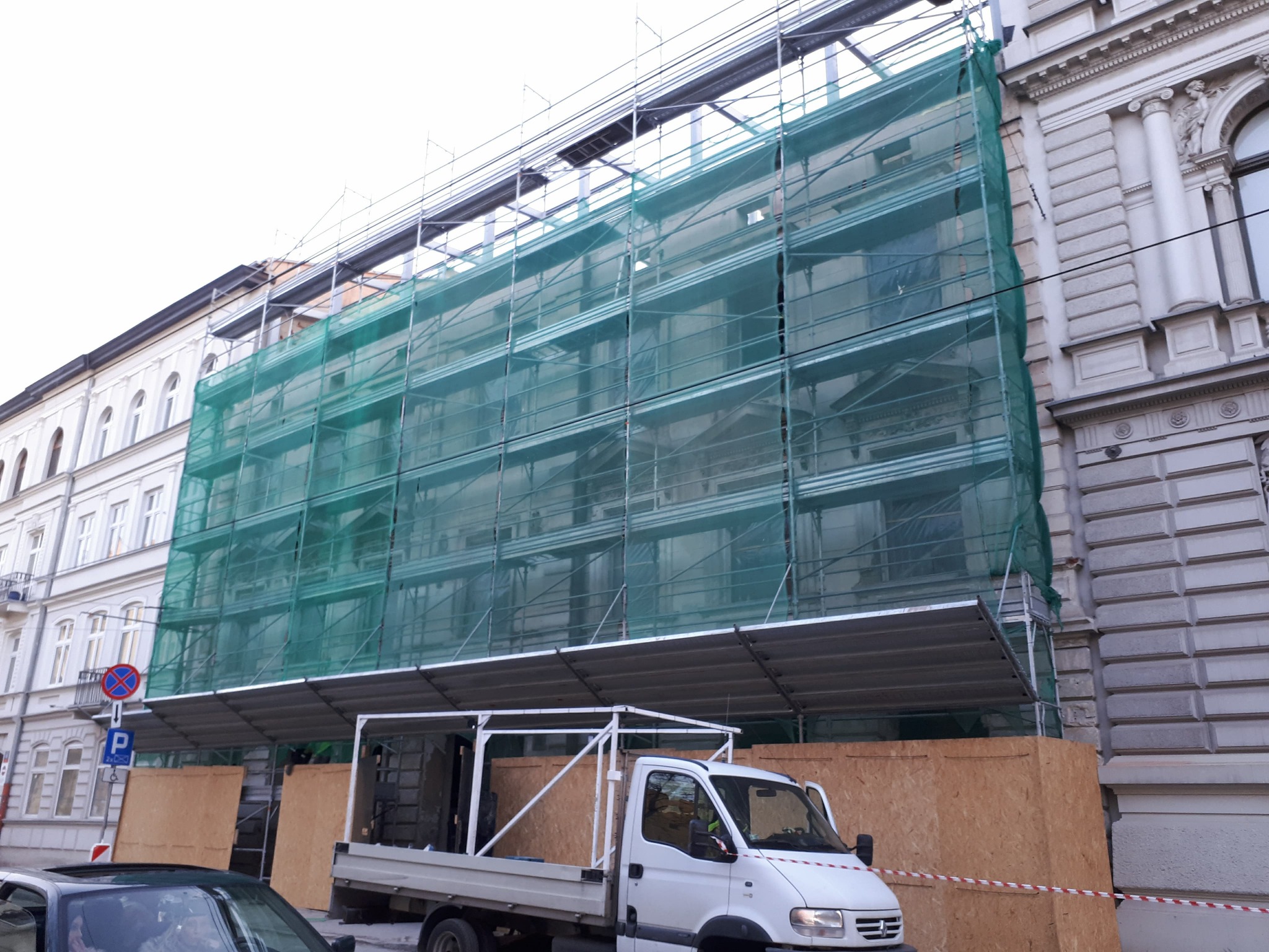 Renowacja elewacji kamienicy w rusztowaniach, zabezpieczona siatką. Na pierwszym planie samochód dostawczy i tymczasowe zabezpieczenia z płyt OSB.