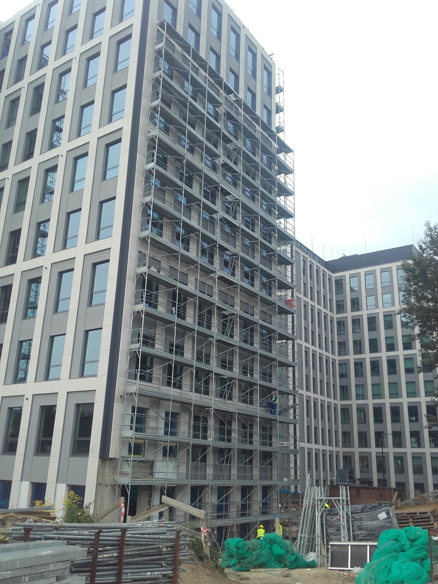 Wielopiętrowy budynek w trakcie prac elewacyjnych, osłonięty rusztowaniami. Widoczne materiały budowlane i pracownicy w odblaskowych kamizelkach. Ujęcie z dołu.