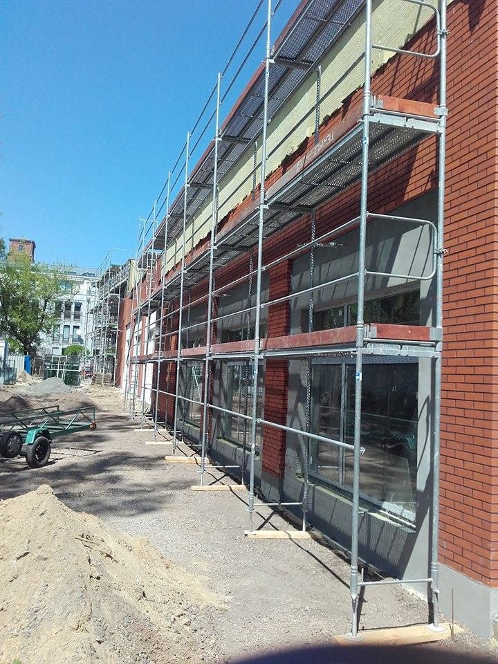 Budynek w trakcie prac elewacyjnych z użyciem rusztowań. Widoczna ceglana ściana i fragmenty ocieplenia. Na pierwszym planie hałda piasku i przyczepka.