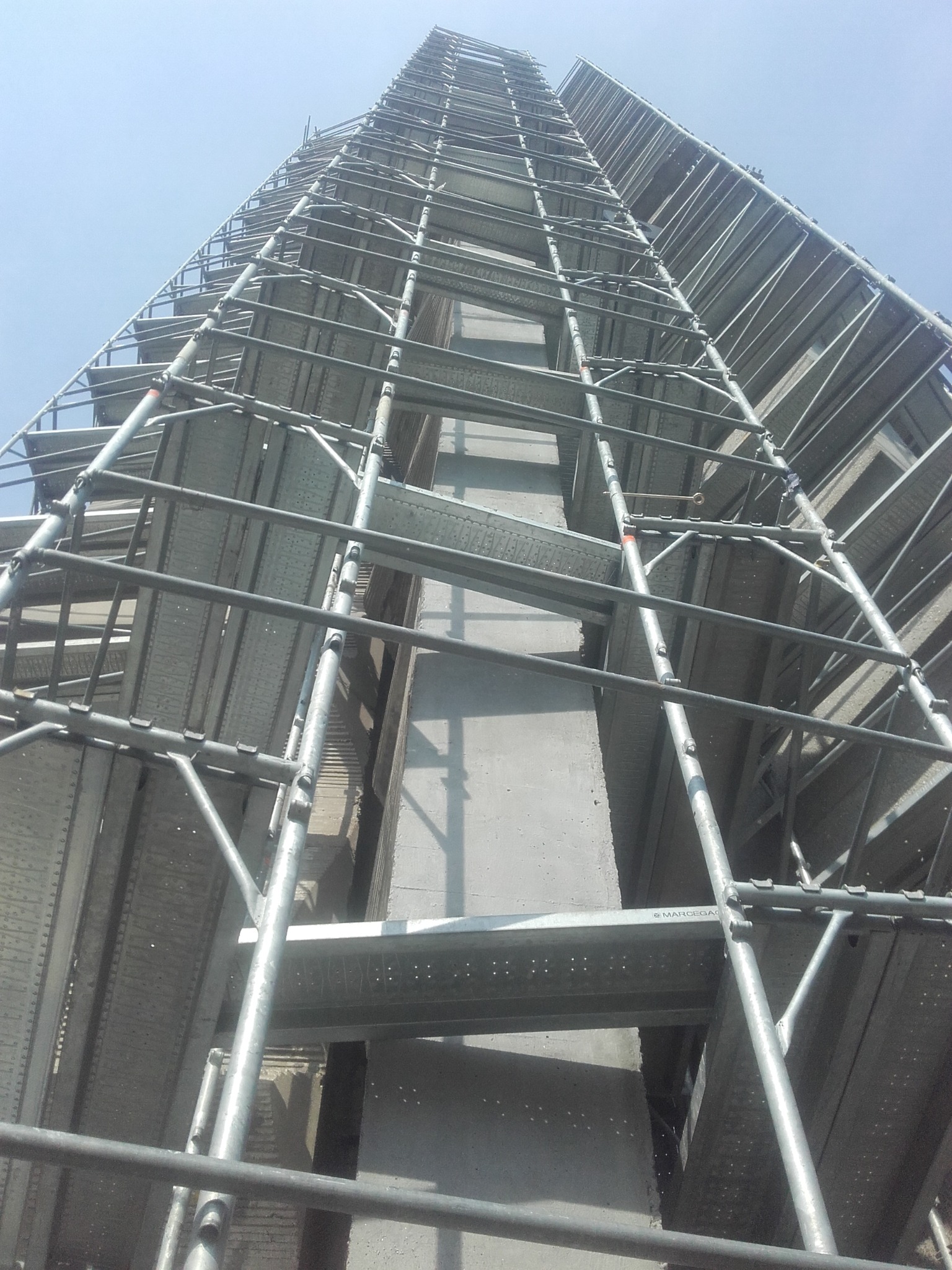 Wysoka konstrukcja rusztowania elewacyjnego, widok z dołu, ukazujący metalowe elementy i fragment fasady budynku w trakcie prac. Jasne niebo w tle.