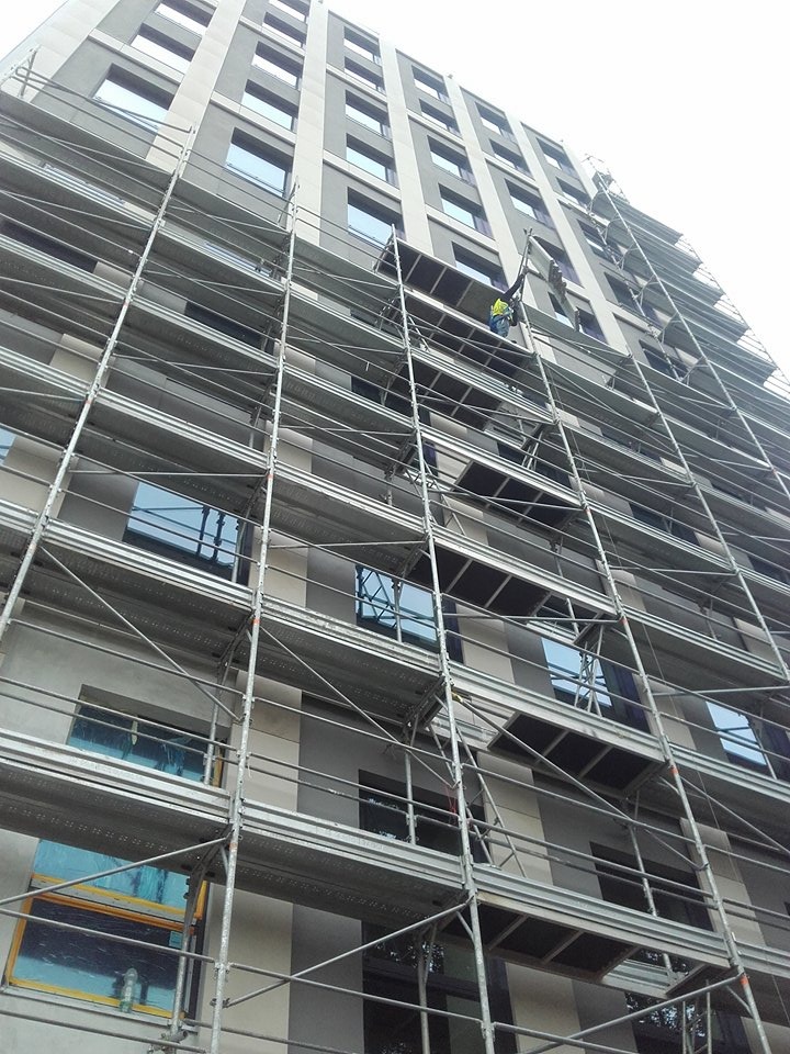 Wysoki budynek w trakcie prac elewacyjnych, osłonięte okna folią, widoczne rusztowania i pracownik w odblaskowej kamizelce na wysokości. Ujęcie z dołu.