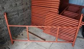 Pomarańczowe, metalowe rusztowanie modułowe na tle szarej ściany i stosu rur. Widoczne elementy konstrukcyjne, złącza i charakterystyczna geometria rusztowania.