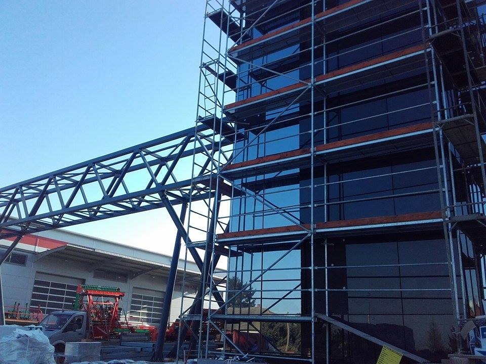 Elewacja budynku biurowego w trakcie montażu, widoczne rusztowania i konstrukcja stalowa łącząca budynki. Nowoczesna architektura z ciemnymi szybami.