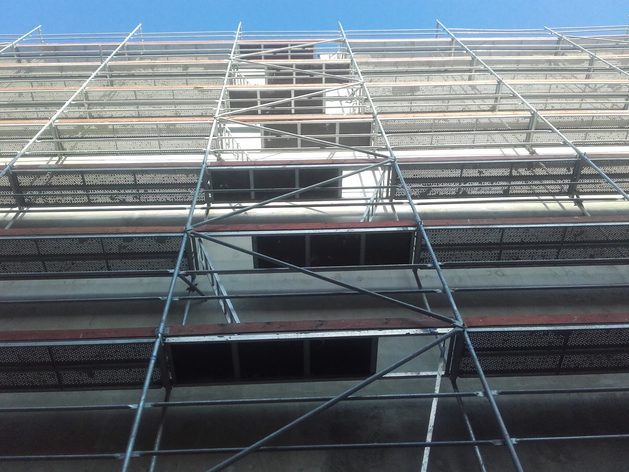 Metalowe rusztowanie na tle ściany budynku w trakcie prac elewacyjnych, widok z dołu, błękitne niebo w tle. Charakterystyczny układ belek i platform.